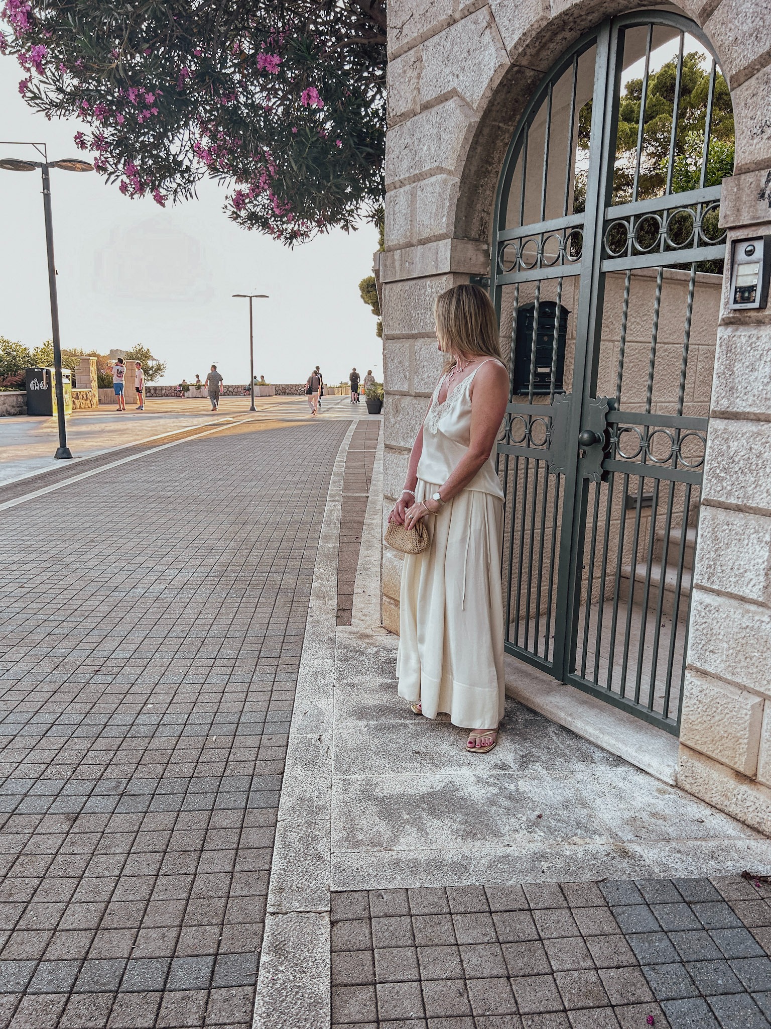 Sarah wearing a cream camisole and wide-leg trousers with a woven bag beside a stone archway